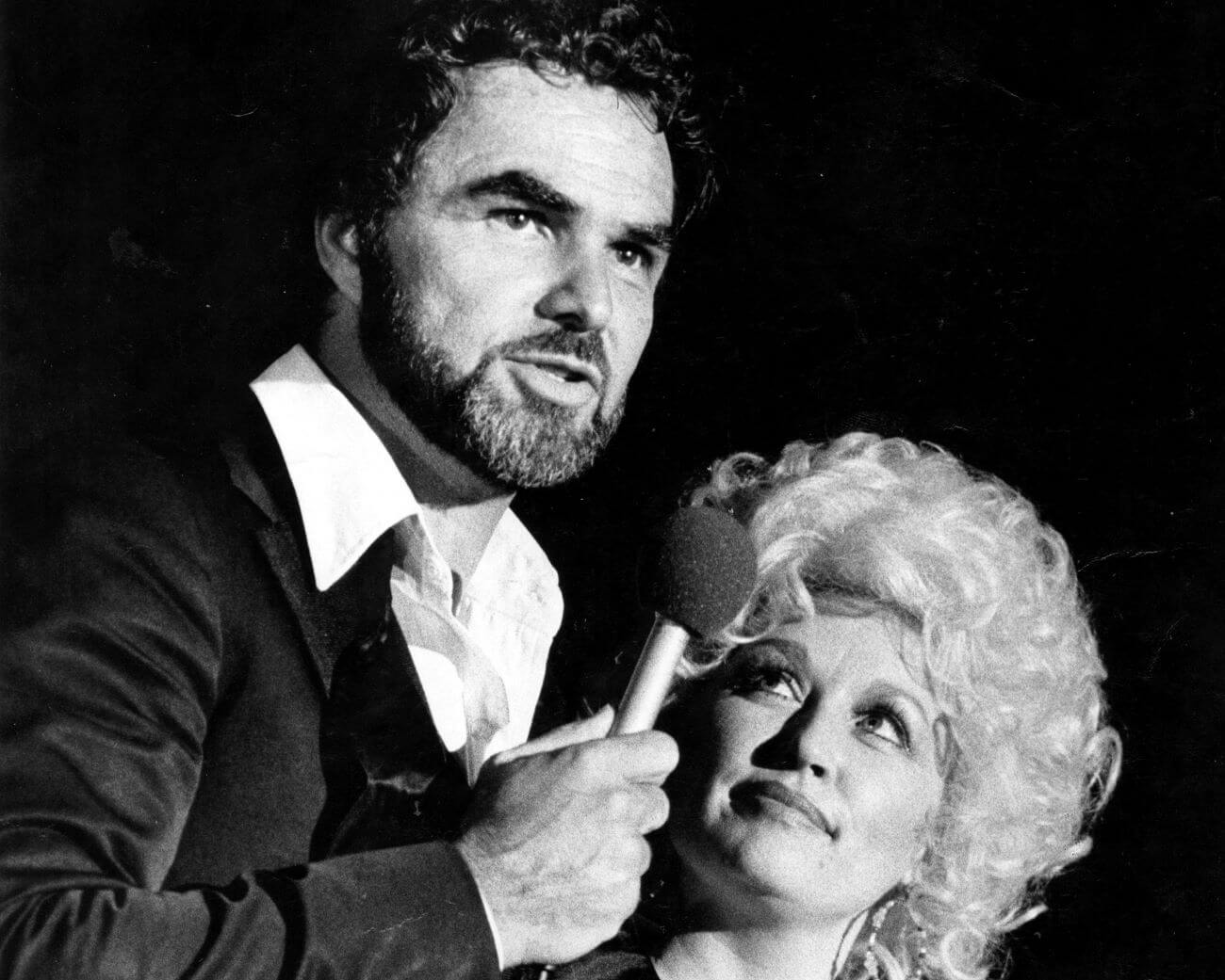 A black and white picture of Burt Reynolds speaking into a microphone and Dolly Parton looking up at him.