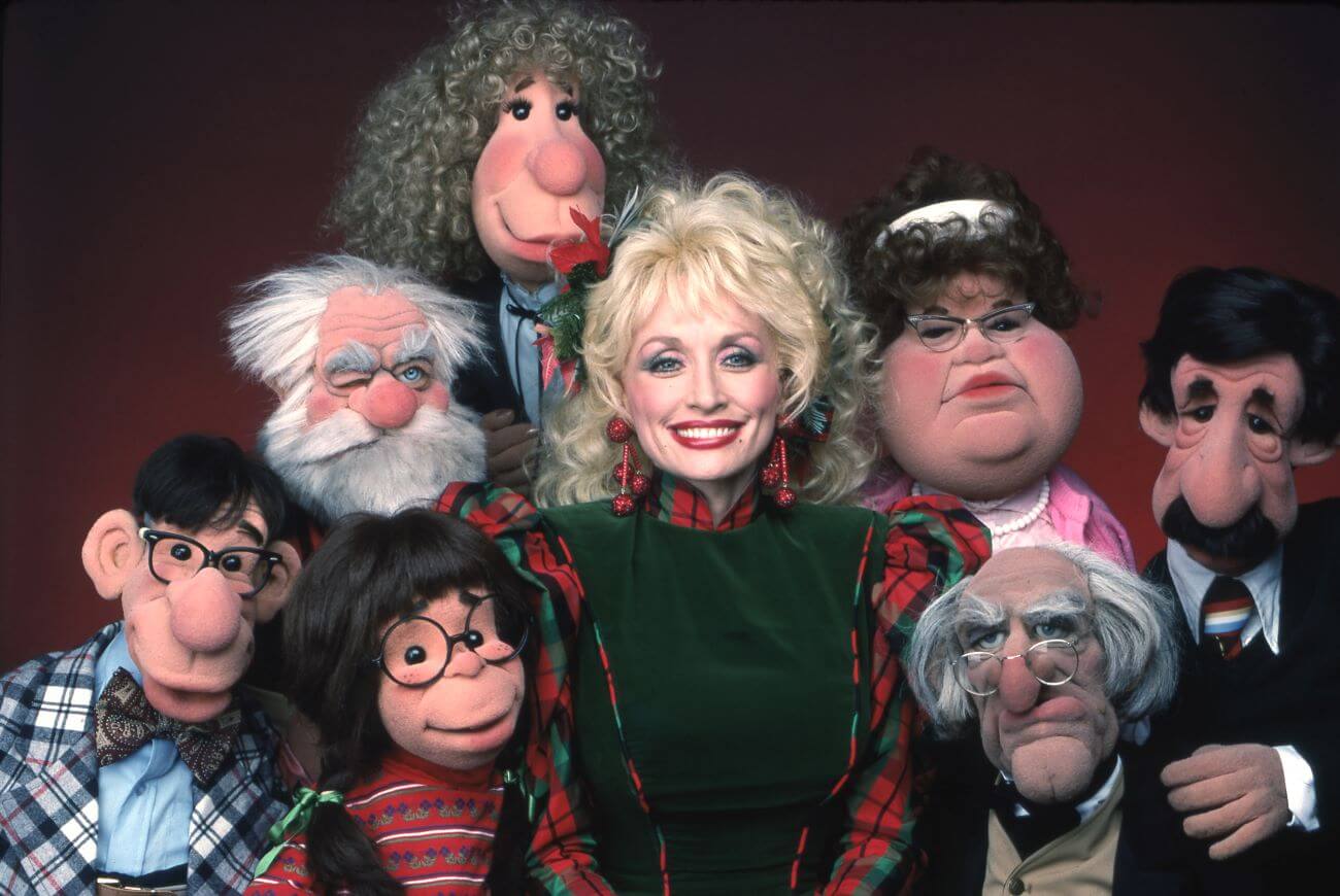 Dolly Parton wears a red plaid dress and poses with puppets.