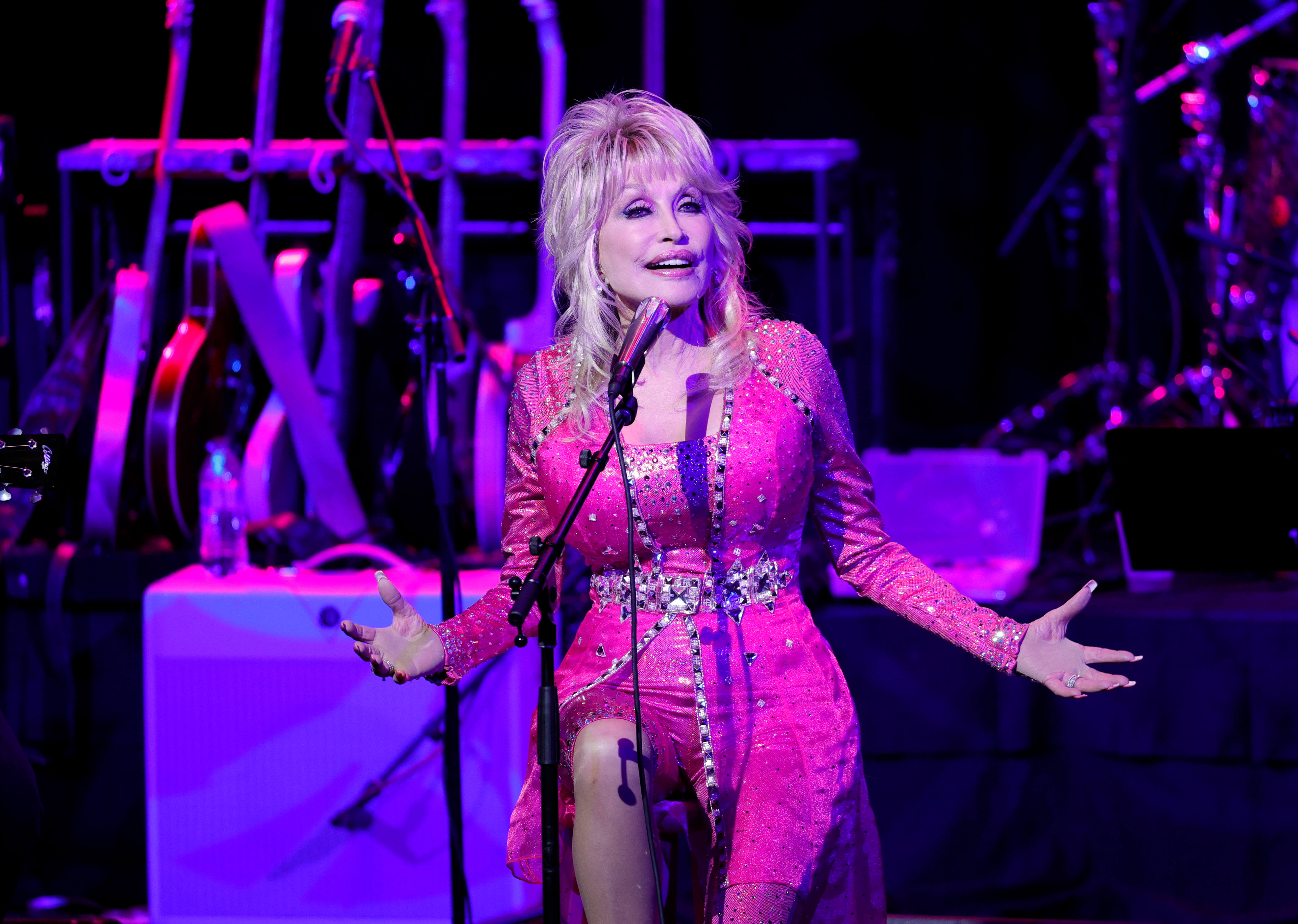 Dolly Parton sings into a microphone in a pink outfit.