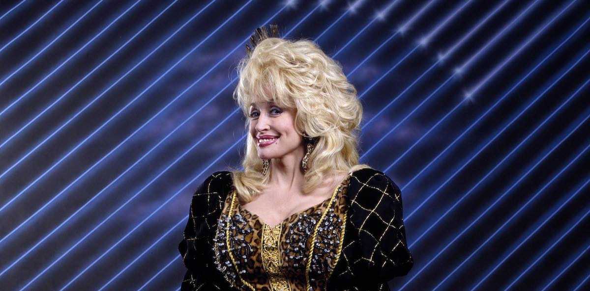 Dolly Parton standing in front of a striped blue background in a promotional image for the unaired sitcom 'Heavens to Betsy'