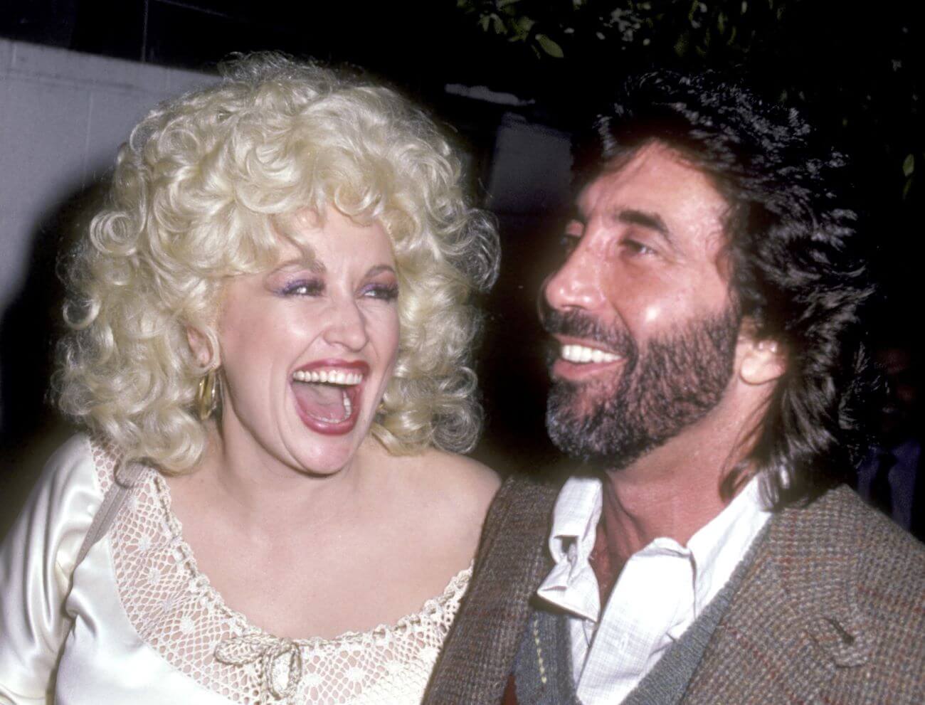 Dolly Parton wears a white dress and laughs while looking at manager Sandy Gallin. He wears a jacket and white shirt.