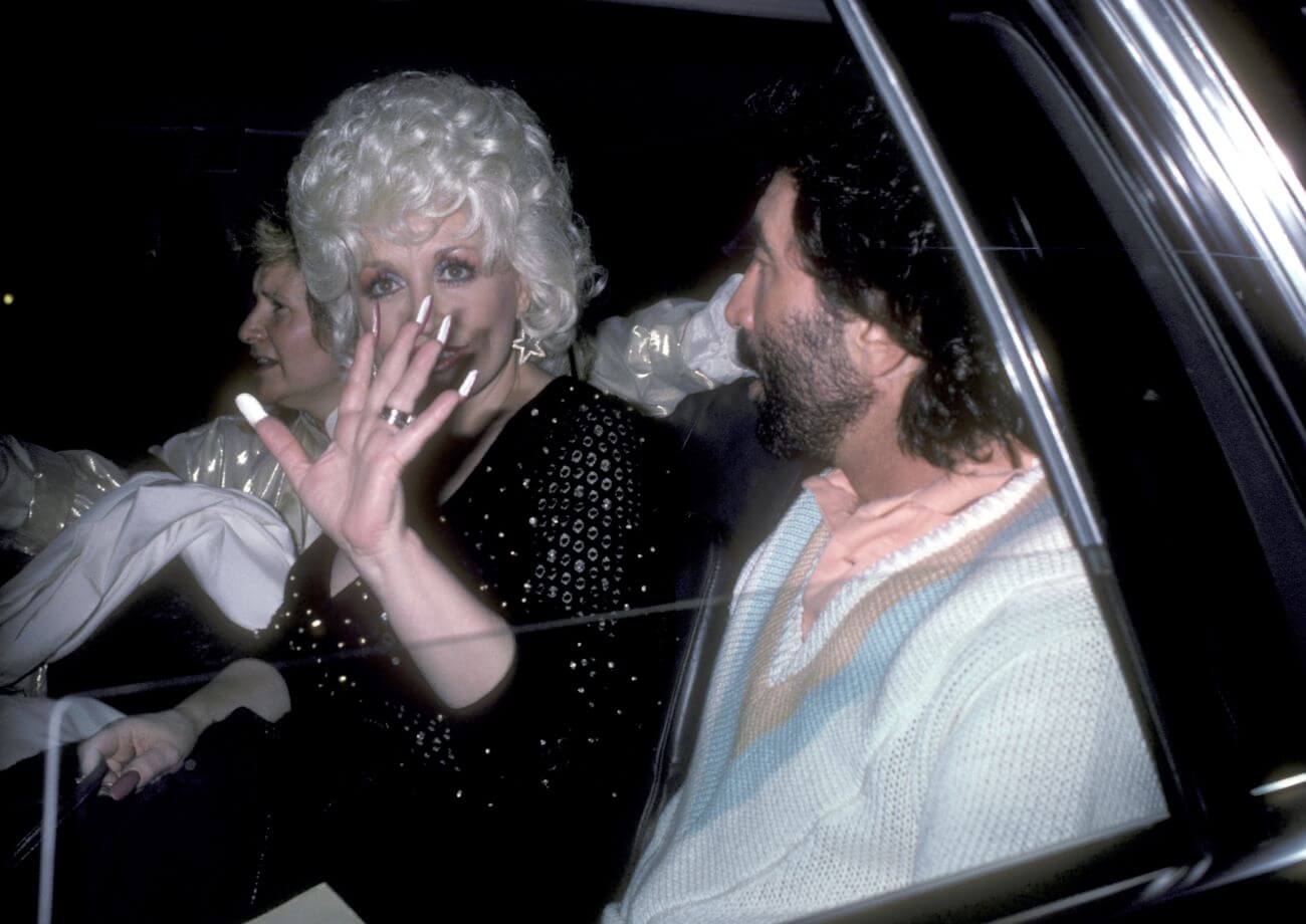 Dolly Parton sits in the backseat of a car with Sandy Gallin. She holds her hand up to the camera.