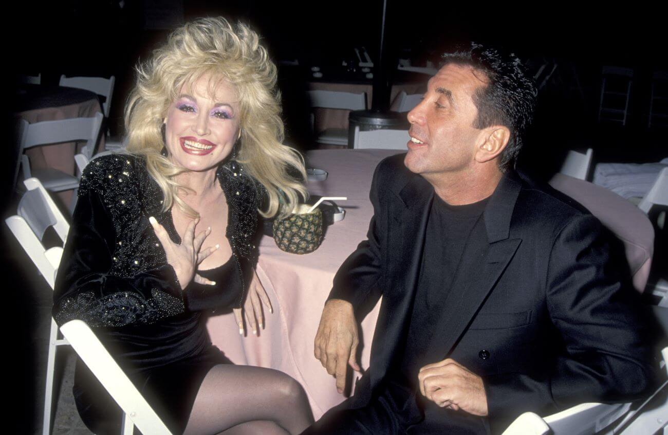 Dolly Parton touches her hand to her chest and smiles while sitting at a table with Sandy Gallin. They both wear black.