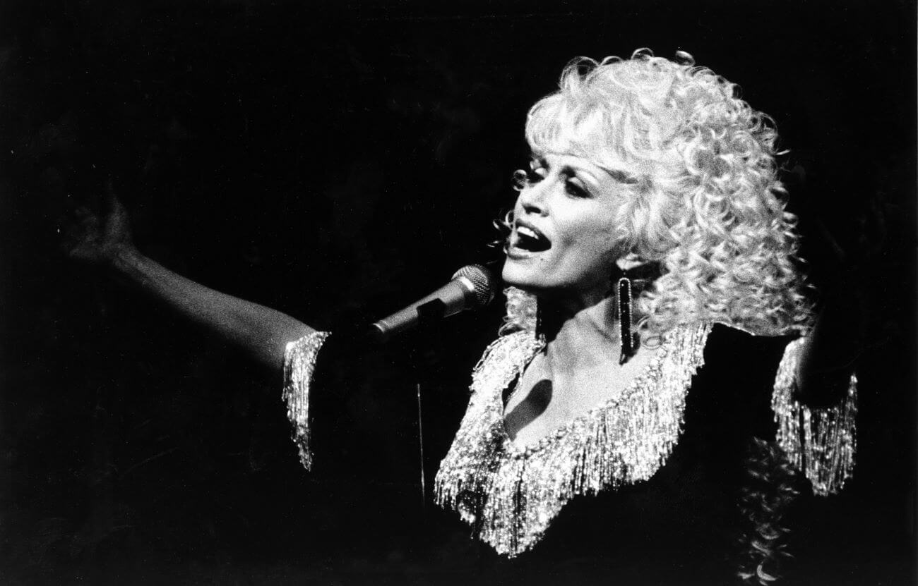 A black and white picture of Dolly Parton singing into a microphone while spreading her arms wide.