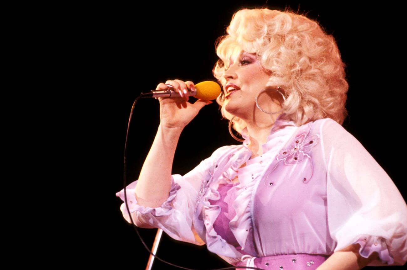 Dolly Parton wears a purple dress and sings into a microphone.