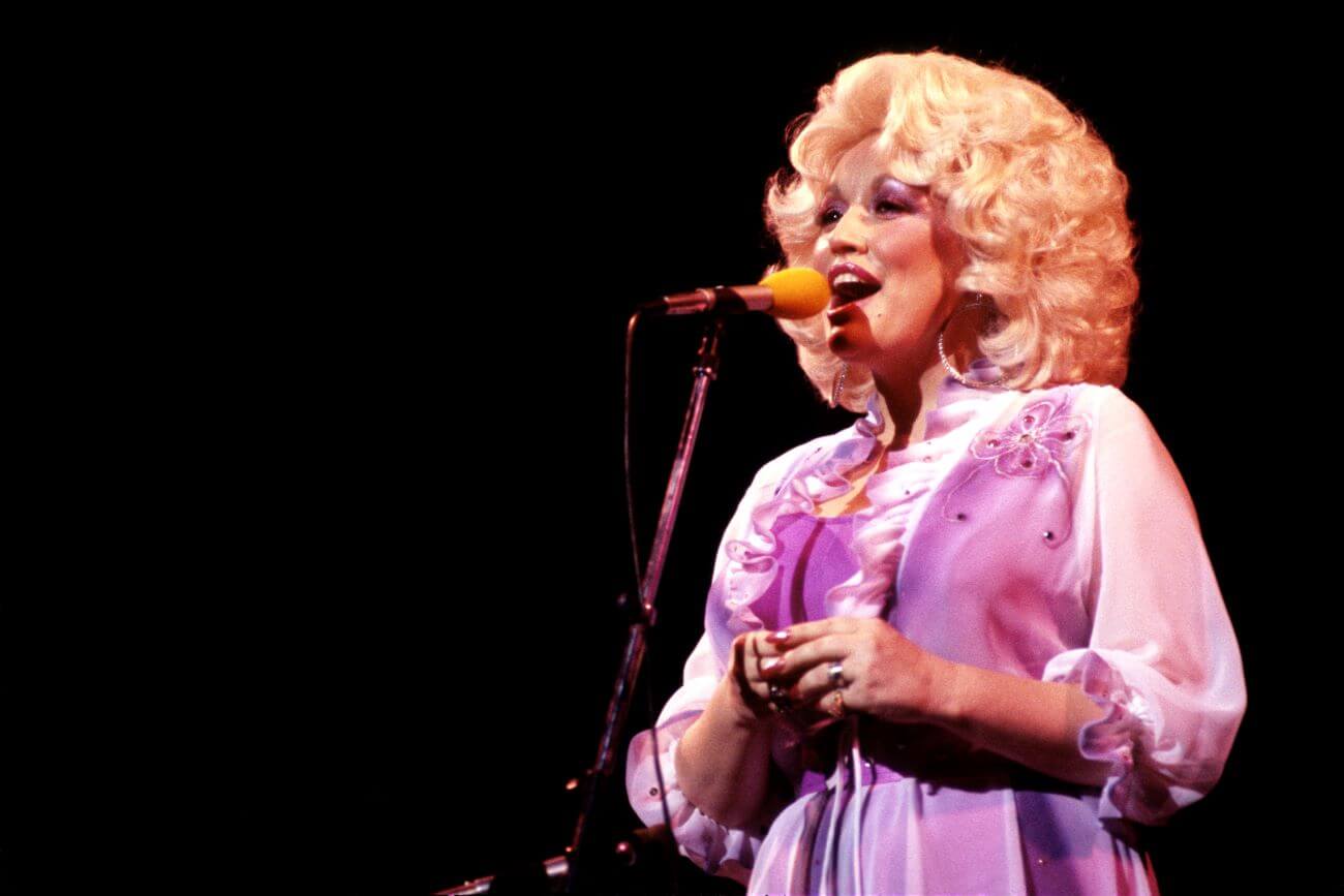 Dolly Parton wears a purple dress and sings into a microphone.