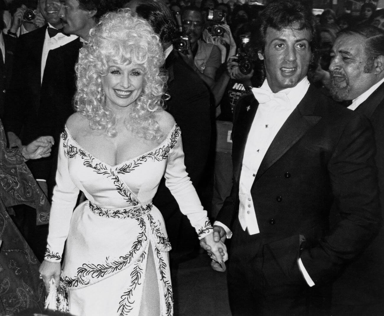 A black and white picture of Dolly Parton and Sylvester Stallone holding hands in a crowd on the set of 'Rhinestone.'