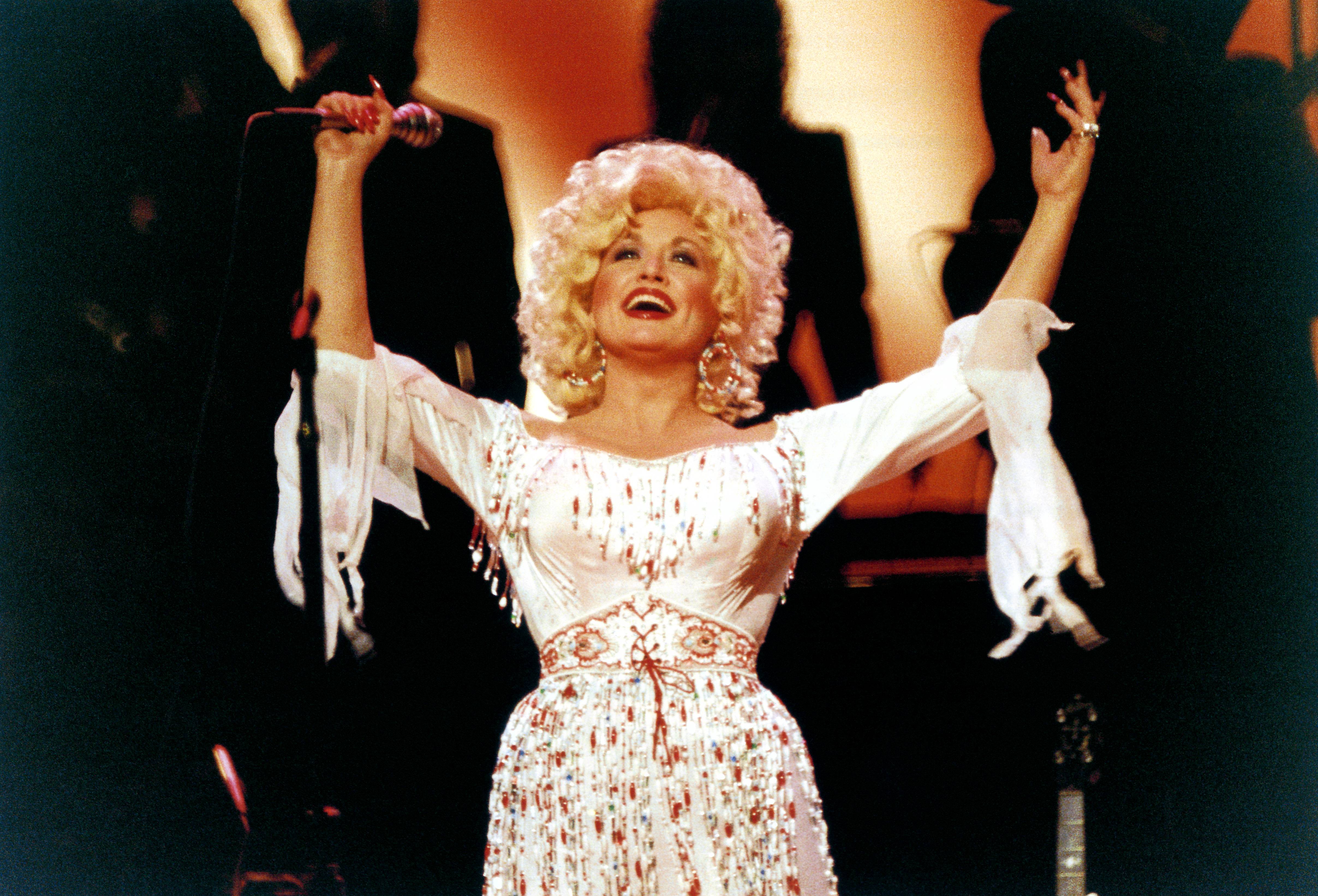 Dolly Parton on stage, singing and raising her arms in the air.
