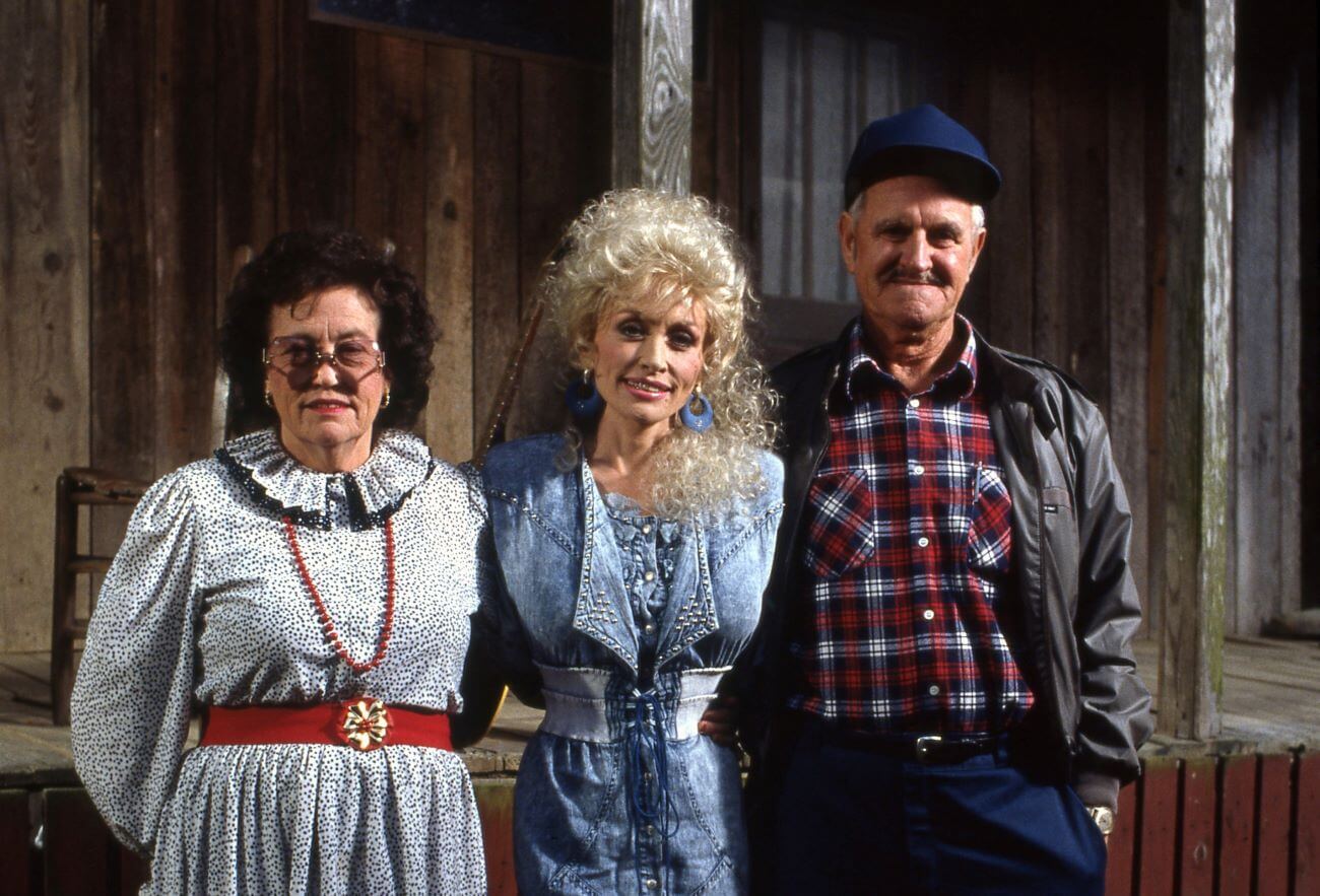 Avie Lee Parton, Dolly Parton, and Lee Parton stand in front of a cabin.