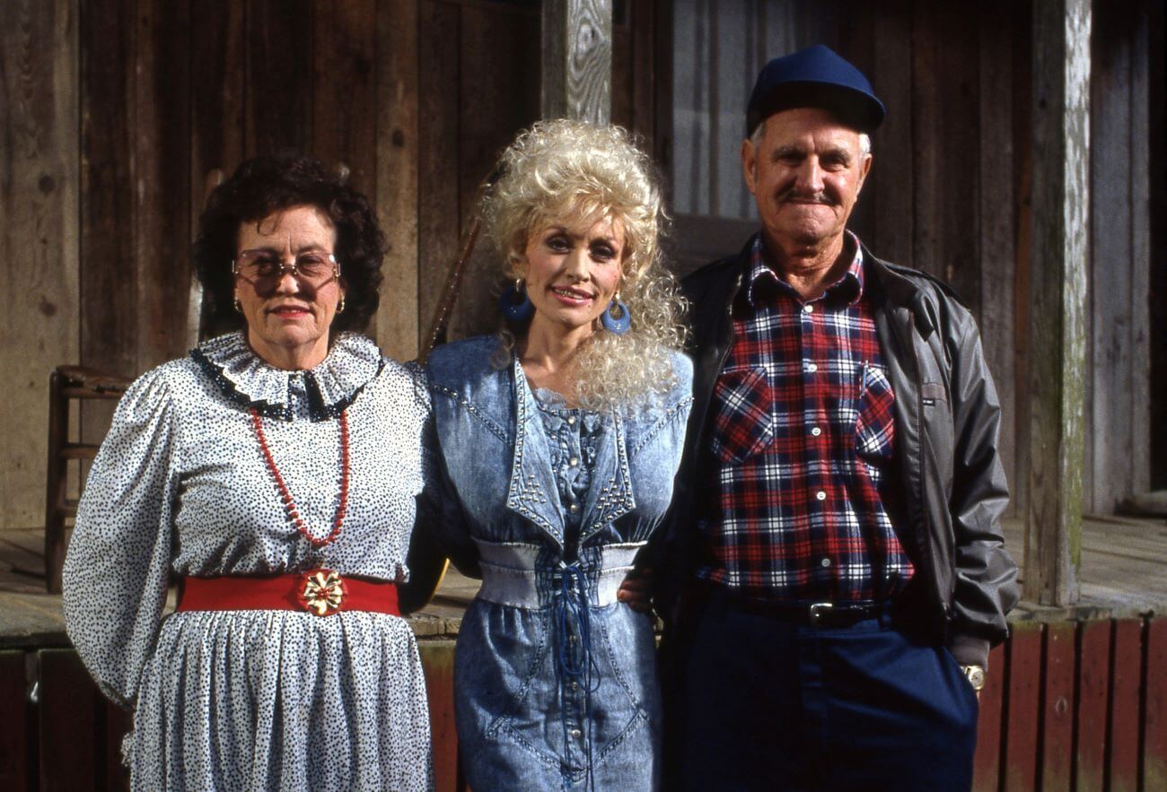 Dolly Parton stands between her mother Avie Lee Parton and her father Robert Lee Parton. They stand in front of a log cabin.