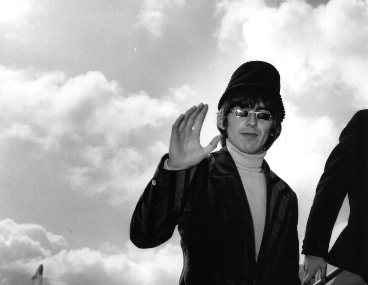 George Harrison wearing rectangular sunglasses, turtleneck, sport coat, and hat while waving to the camera as he boards a plane.