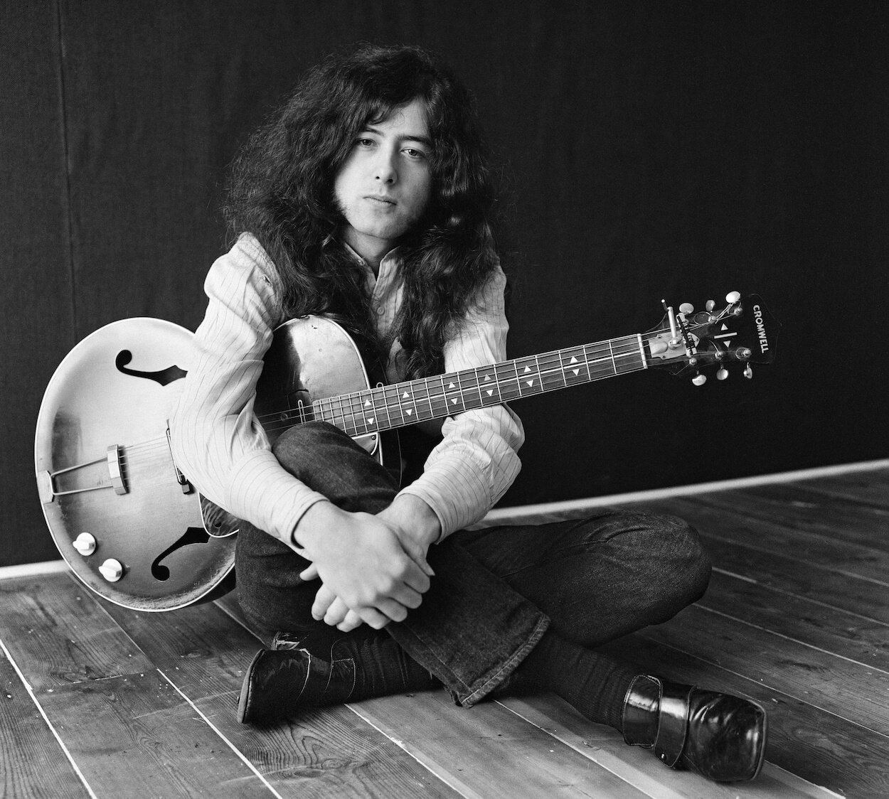 Jimmy Page cradling a guitar and sitting on a wooden floor in his house.