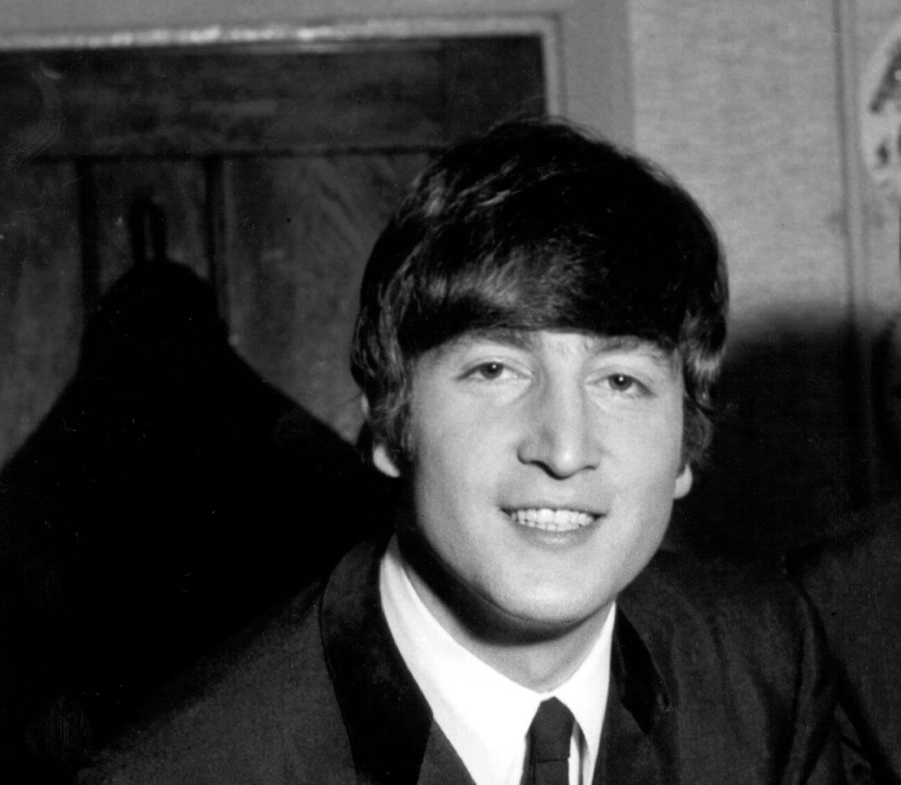A black and white picture of John Lennon wearing a suit and tie. He stands in a doorway.