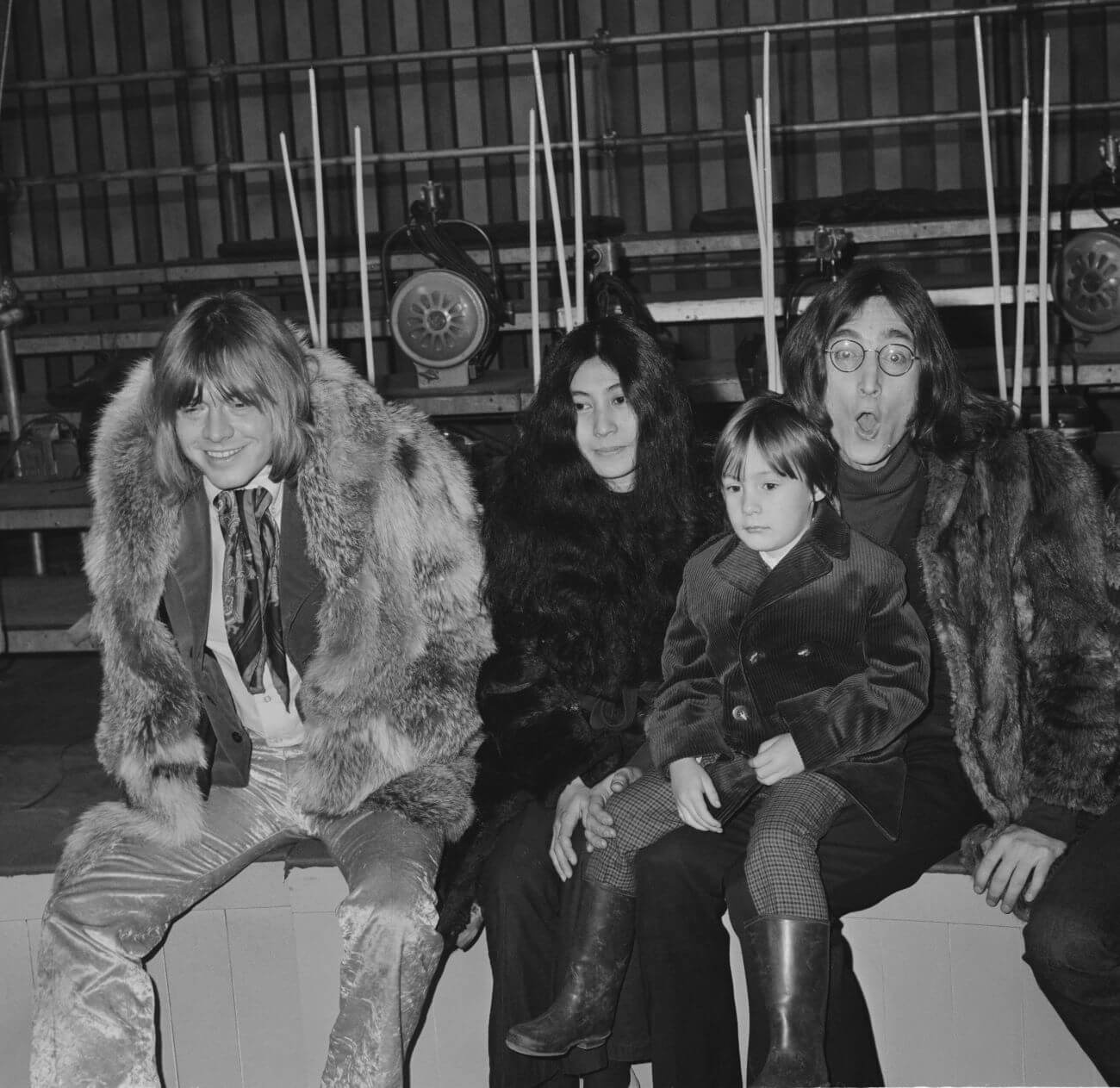 A black and white picture of Brian Jones, Yoko Ono, Julian Lennon, and John Lennon sitting on a bench together. Jones and Lennon wear fur coats.