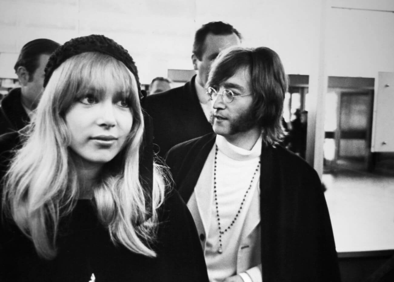 A black and white picture of Pattie Boyd wearing a hat and walking slightly ahead of John Lennon.