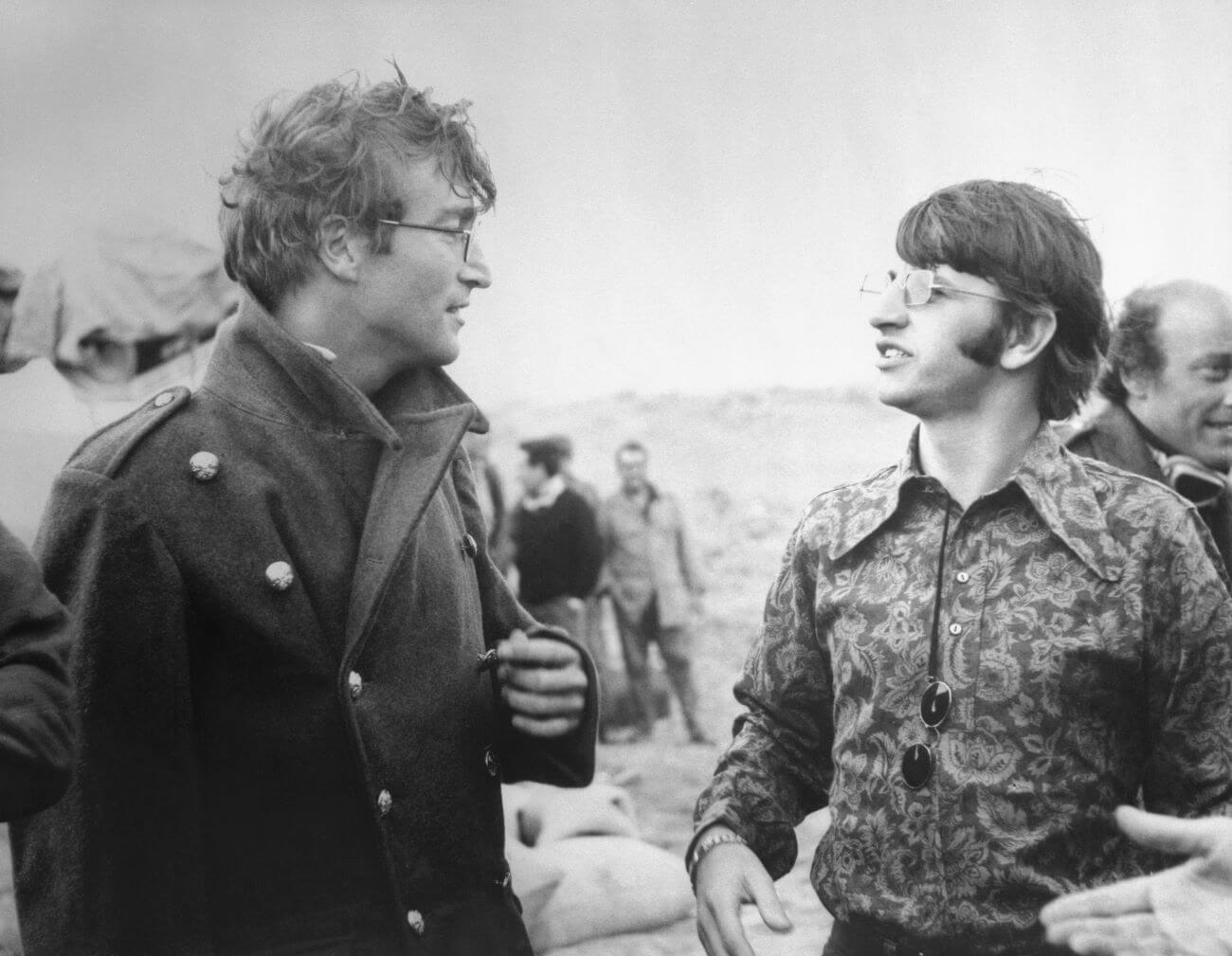 A black and white picture of John Lennon and Ringo Starr standing outside and speaking to each other.