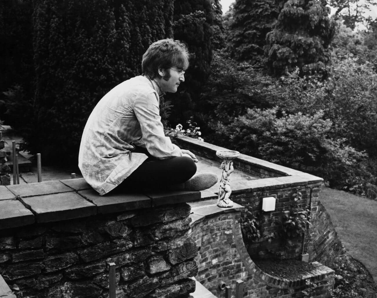 A black and white picture of John Lennon sitting cross-legged on a stone wall.