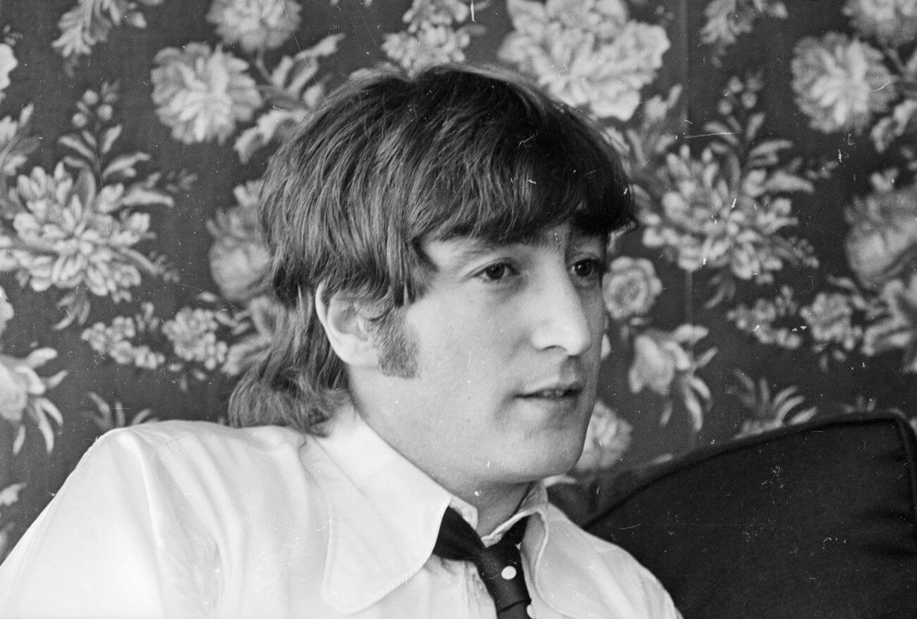 A black and white picture of John Lennon wearing a tie and sitting in front of a wall with floral wallpaper.