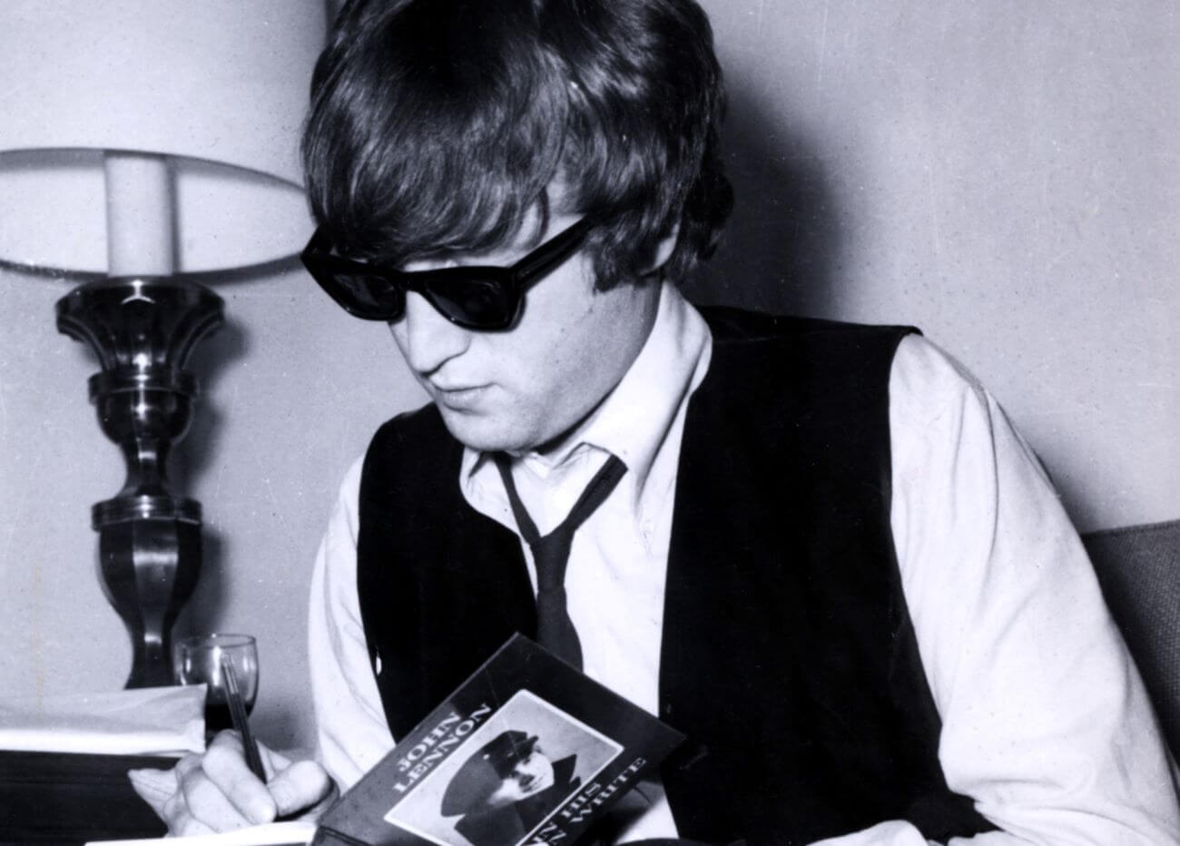 A black and white picture of John Lennon wearing sunglasses and signing a copy of his book "In His Own Write."