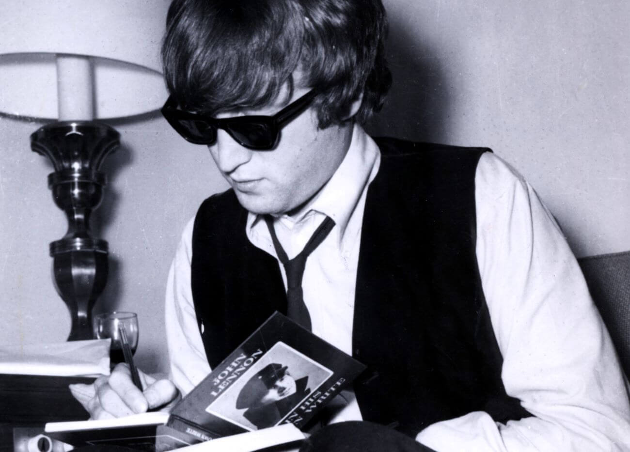 A black and white picture of John Lennon wearing sunglasses and signing a copy of his book "In His Own Write."