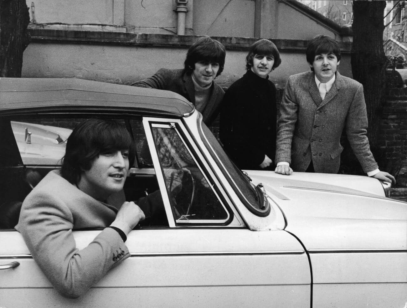 A black and white picture of John Lennon sitting in a car. George Harrison, Ringo Starr, and Paul McCartney stand on the opposite side of the car.