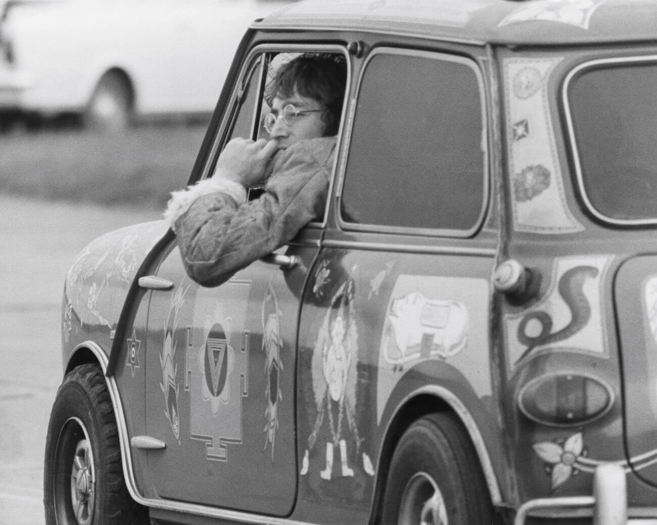 A black and white picture of John Lennon leaning out the window of a car. He sits in the front seat.