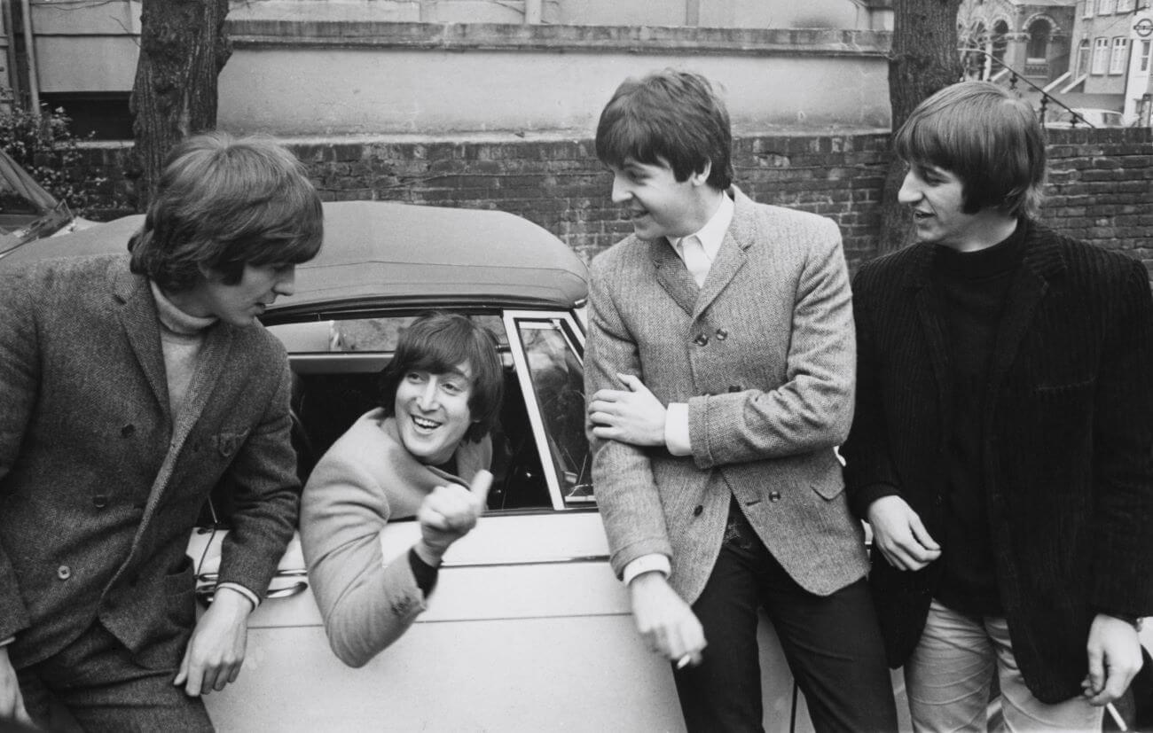 A black and white picture of John Lennon leaning out the window of a car while George Harrison, Paul McCartney, and Ringo Starr stand outside.