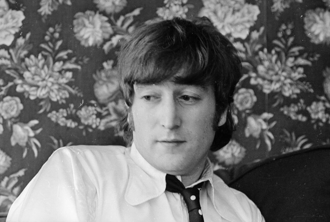 A black and white picture of John Lennon wearing a button down shirt and tie. He sits in front of a wall with floral wallpaper.