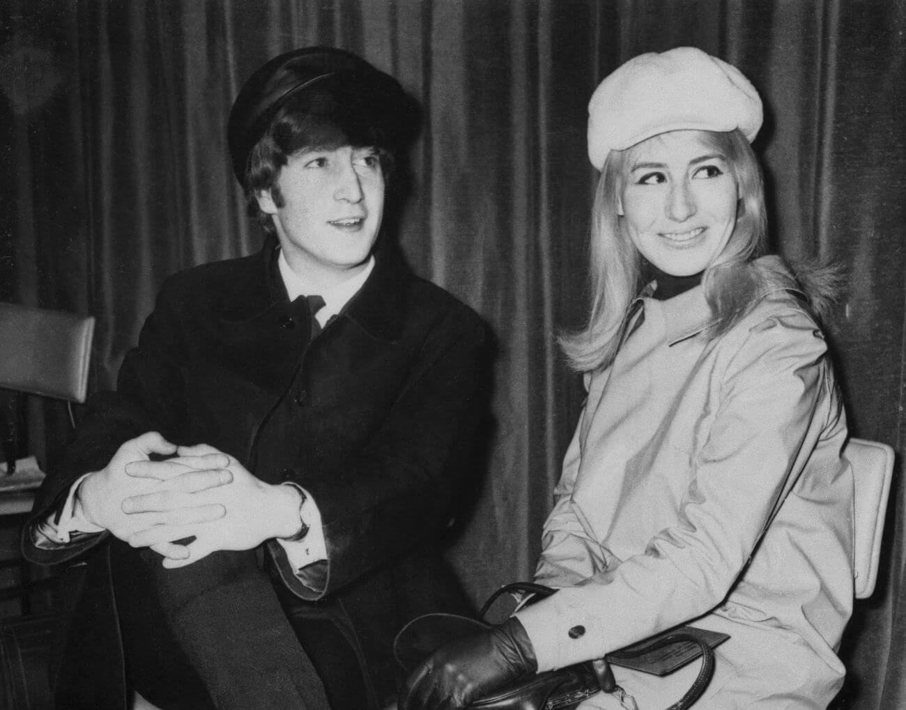 A black and white picture of John Lennon sitting with his hands clasped and his wife Cynthia Lennon wearing a hat and coat.