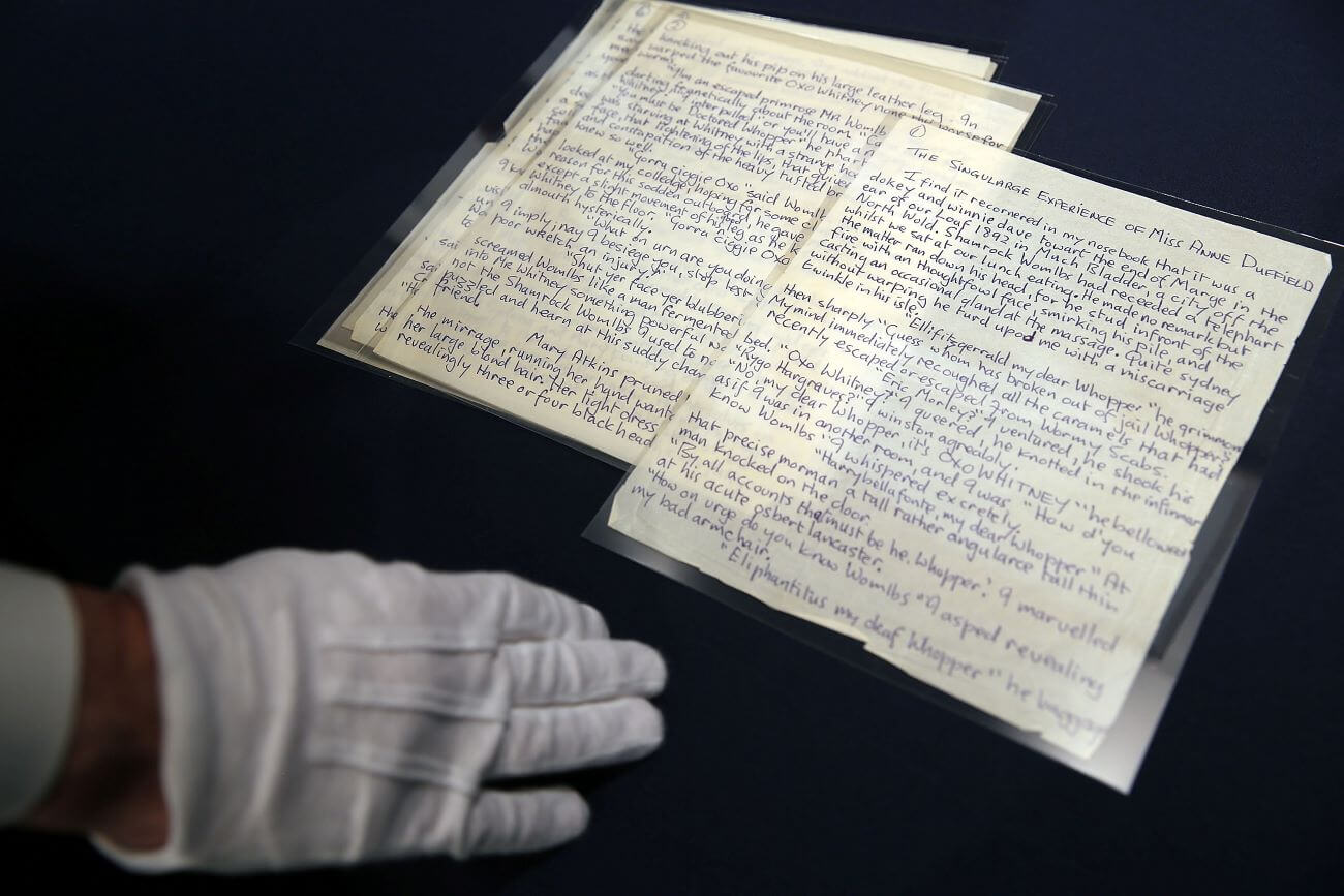 Stacked papers of John Lennon's writing  are on a black surface. A person's white gloved hand rests near the documents. 