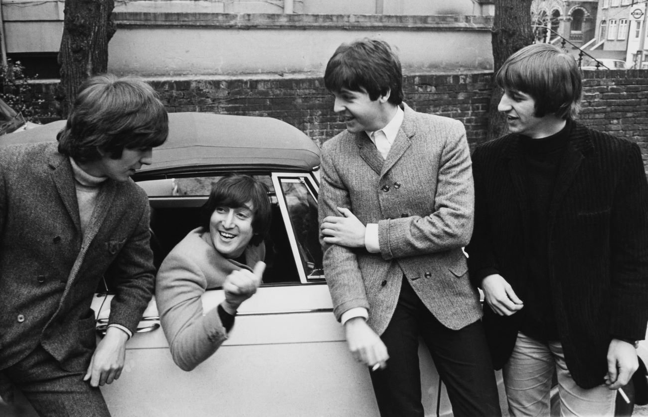 A black and white picture of John Lennon sitting behind the wheel of a car while George Harrison, Paul McCartney, and Ringo Starr stand outside.