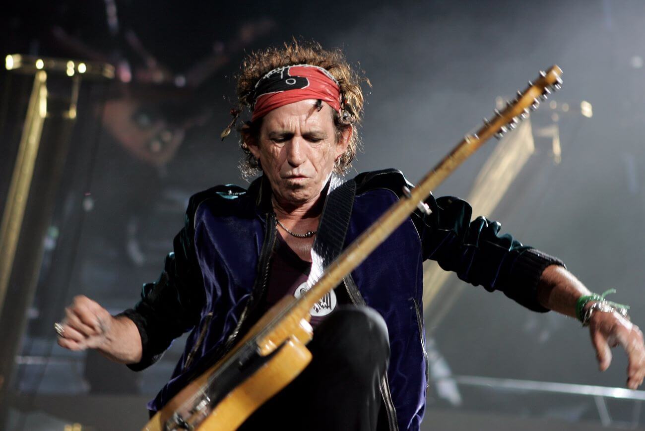 Keith Richards wears a red bandana around his head and takes his hands from his guitar, which is on a strap around his body.