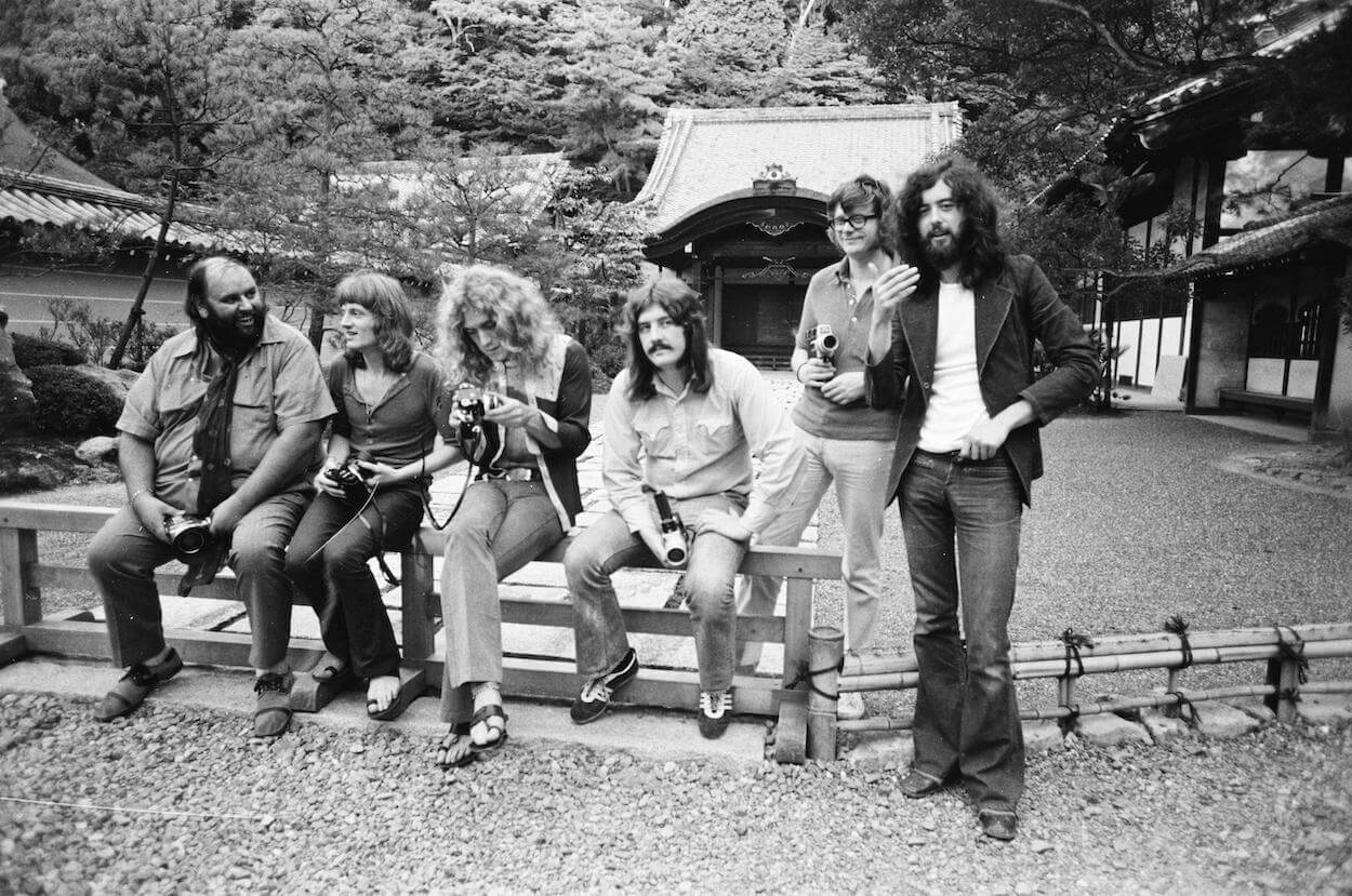 Peter Grant (left) and Led Zeppelin members John Paul Jones, Robert Plant, and John Bonham sitting on a fence while Jimmy Page (far right) stands.