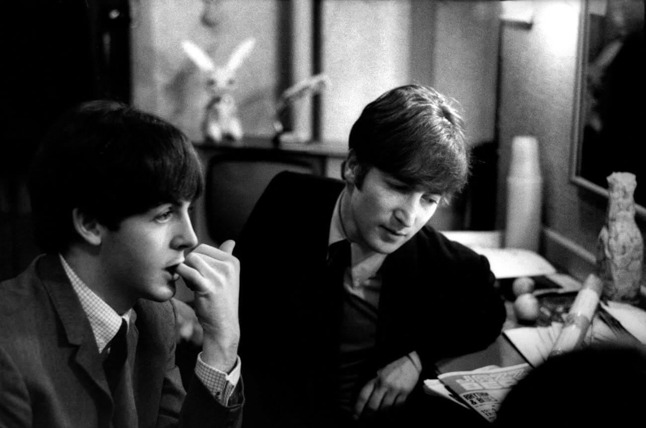 A black and white picture of Paul McCartney and John Lennon sitting at a desk together. McCartney bites his finger.