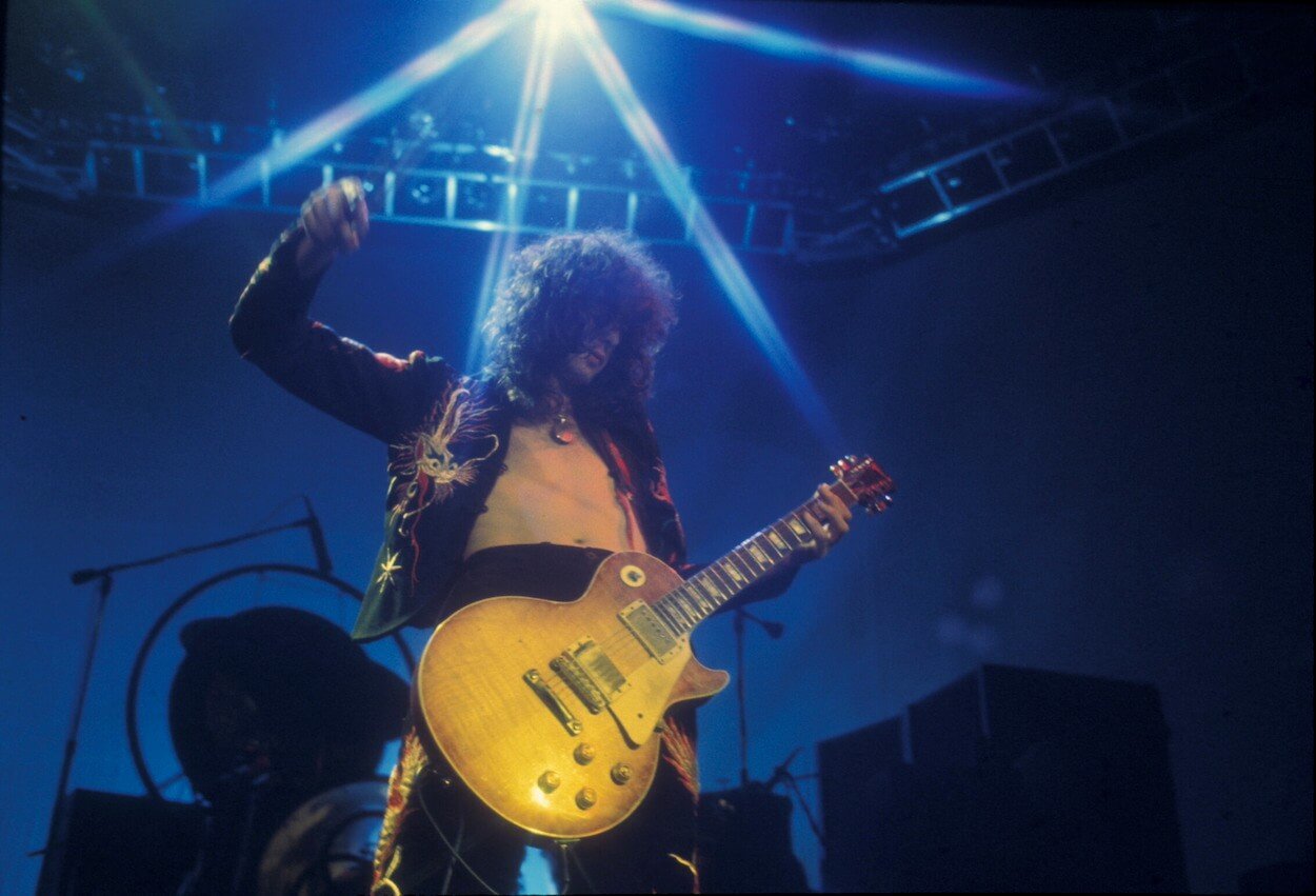 Jimmy Page raising his right hand in the air while playing a concert in 1975.