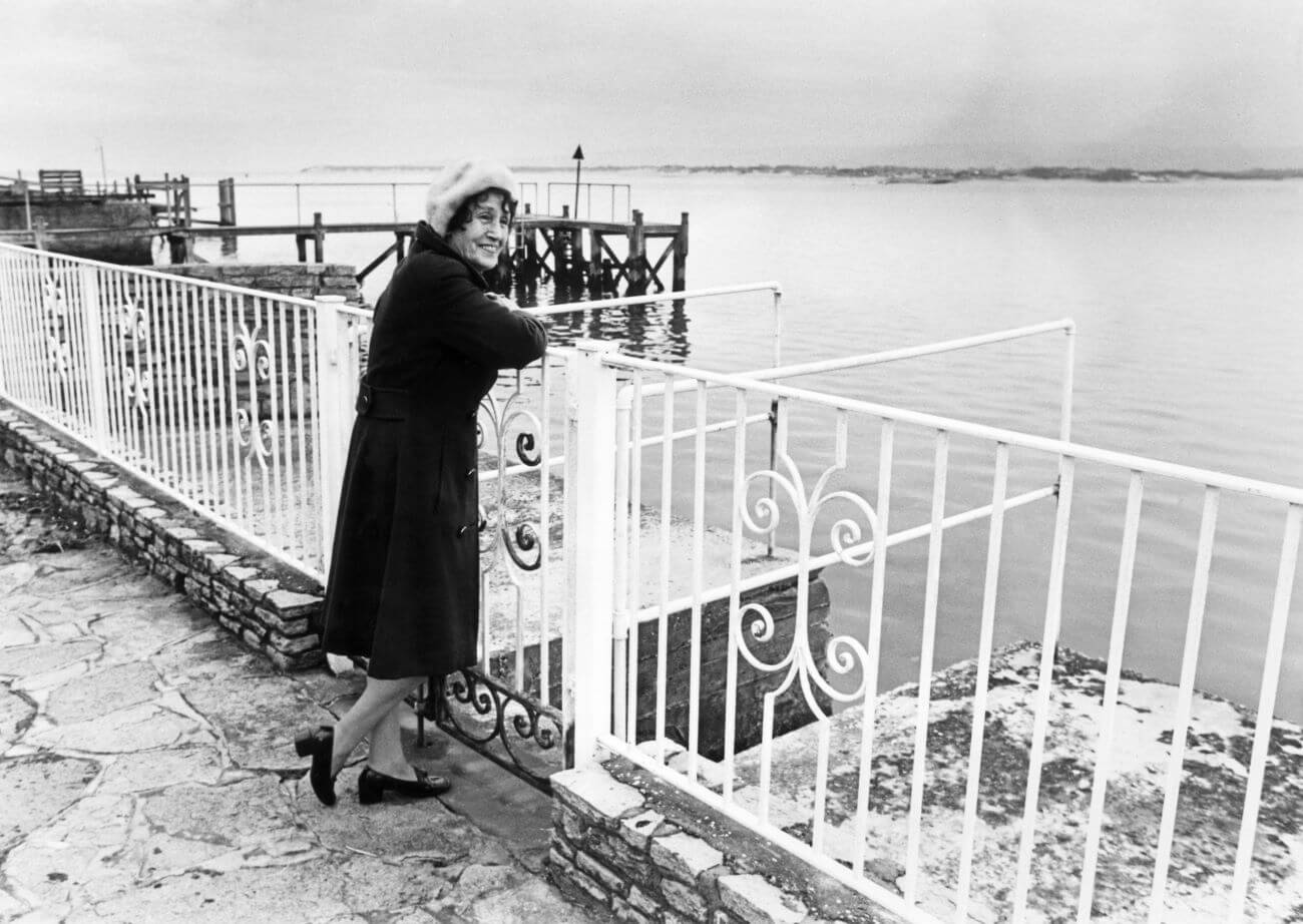A black and white picture of John Lennon's aunt Mimi Smith standing against a fence overlooking the water.