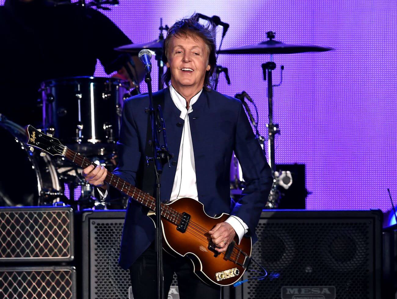 Paul McCartney holds a guitar while standing in front of a drum set. 