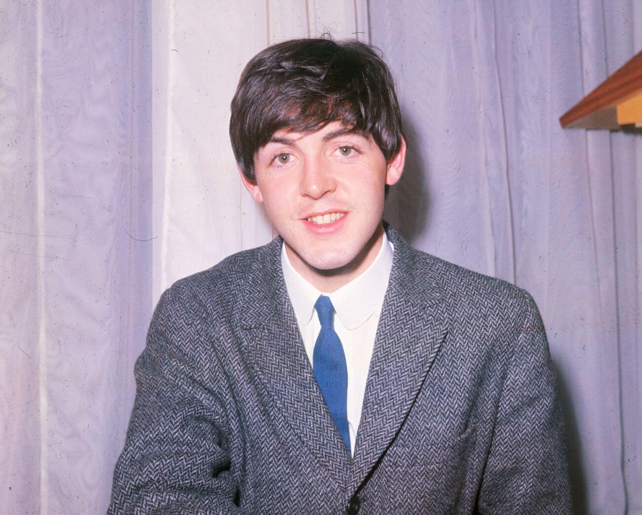 Paul McCartney wears a tweed jacket and a tie and sits in front of a purple curtain.