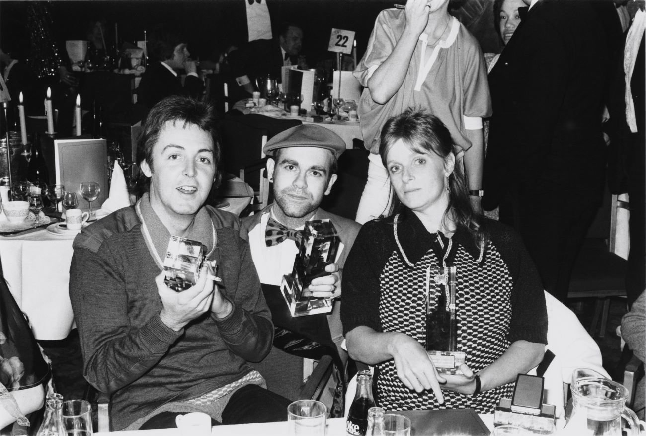 A black and white picture of Paul McCartney, Elton John, and Linda McCartney sitting at a dinner table holding awards.