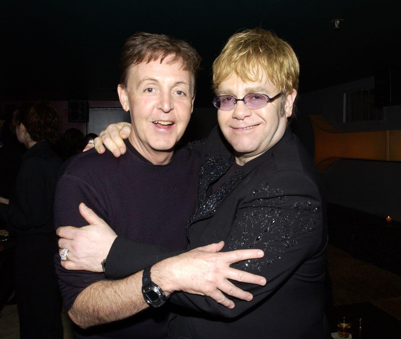 Paul McCartney and Elton John both wear black and embrace each other.