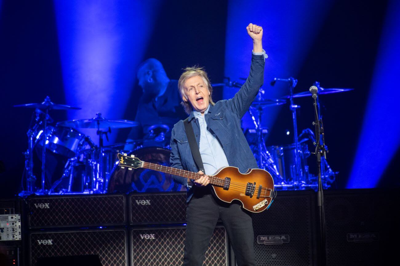 Paul McCartney holds a guitar with one hand and lifts his other fist in the air while on a stage.
