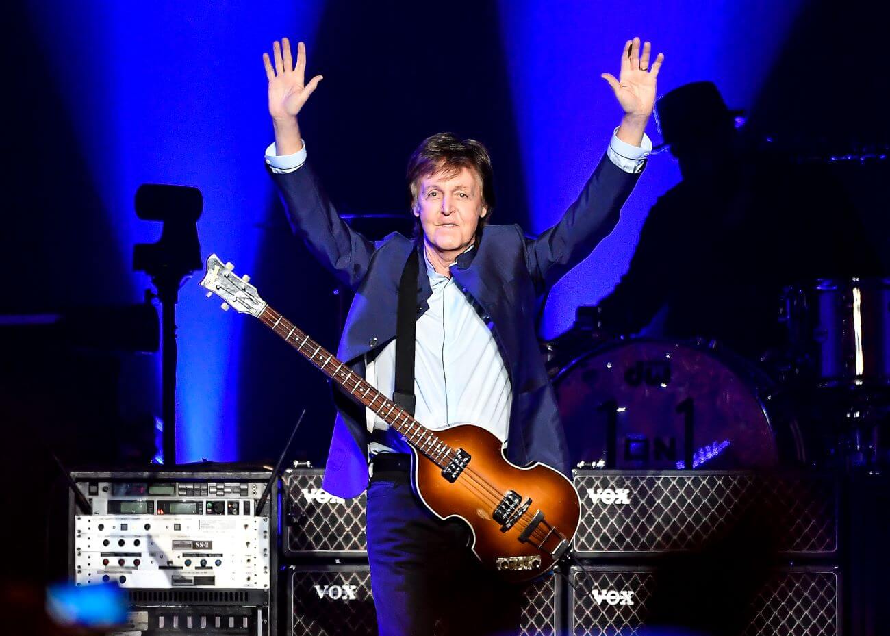 Paul McCartney has a guitar on a strap around his neck and holds his arms in the air.
