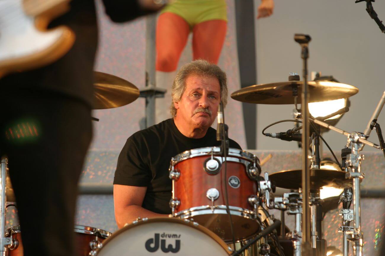 Pete Best wears a black shirt and sits at a drum set. 