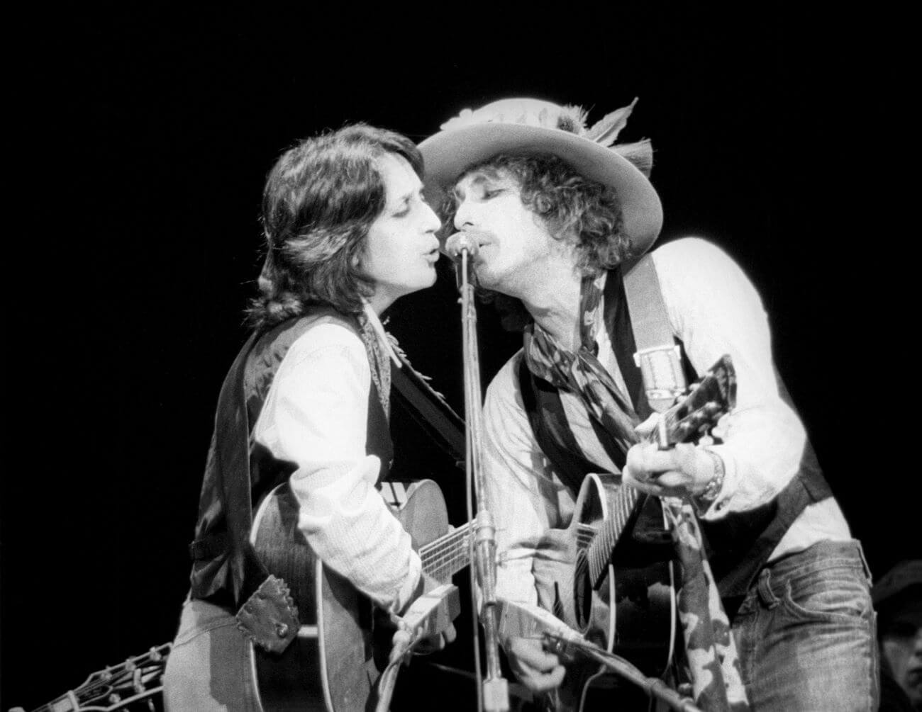 A black and white picture of Joan Baez and Bob Dylan playing guitars and singing into the same microphone. Dylan wears a hat with a feather in it.