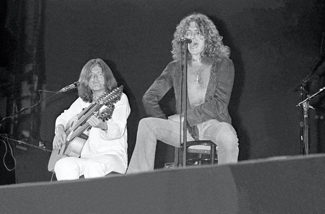 John Paul Jones (left) wearing white and playing triple-necked acoustic guitar while Robert Plant sings during a 1975 Led Zeppelin concert.