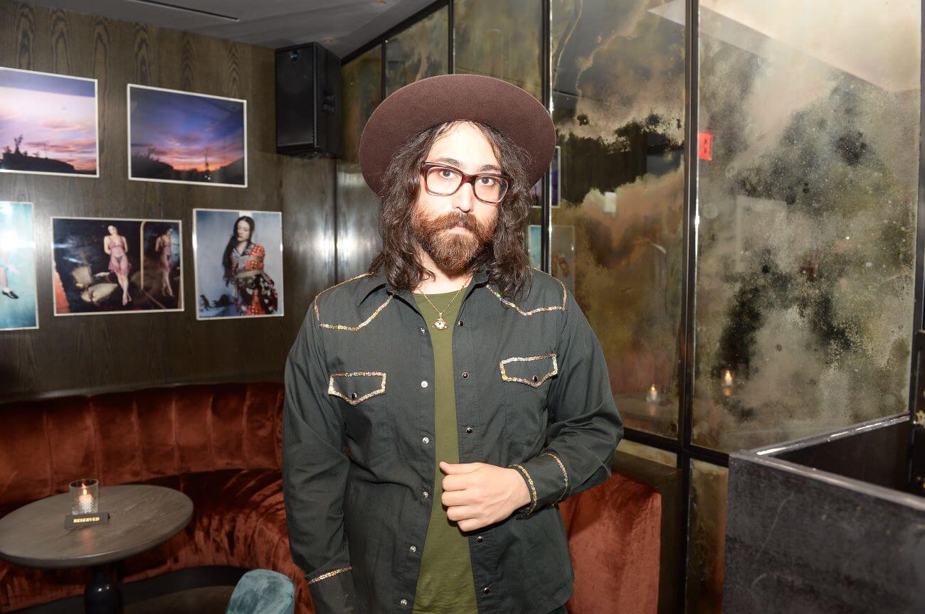 Sean Lennon stands in front of a booth wearing a hat and glasses.