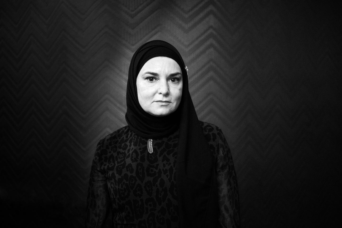 A black and white picture of Sinead O'Connor wearing a hijab in front of a wall with zig-zag wallpaper.