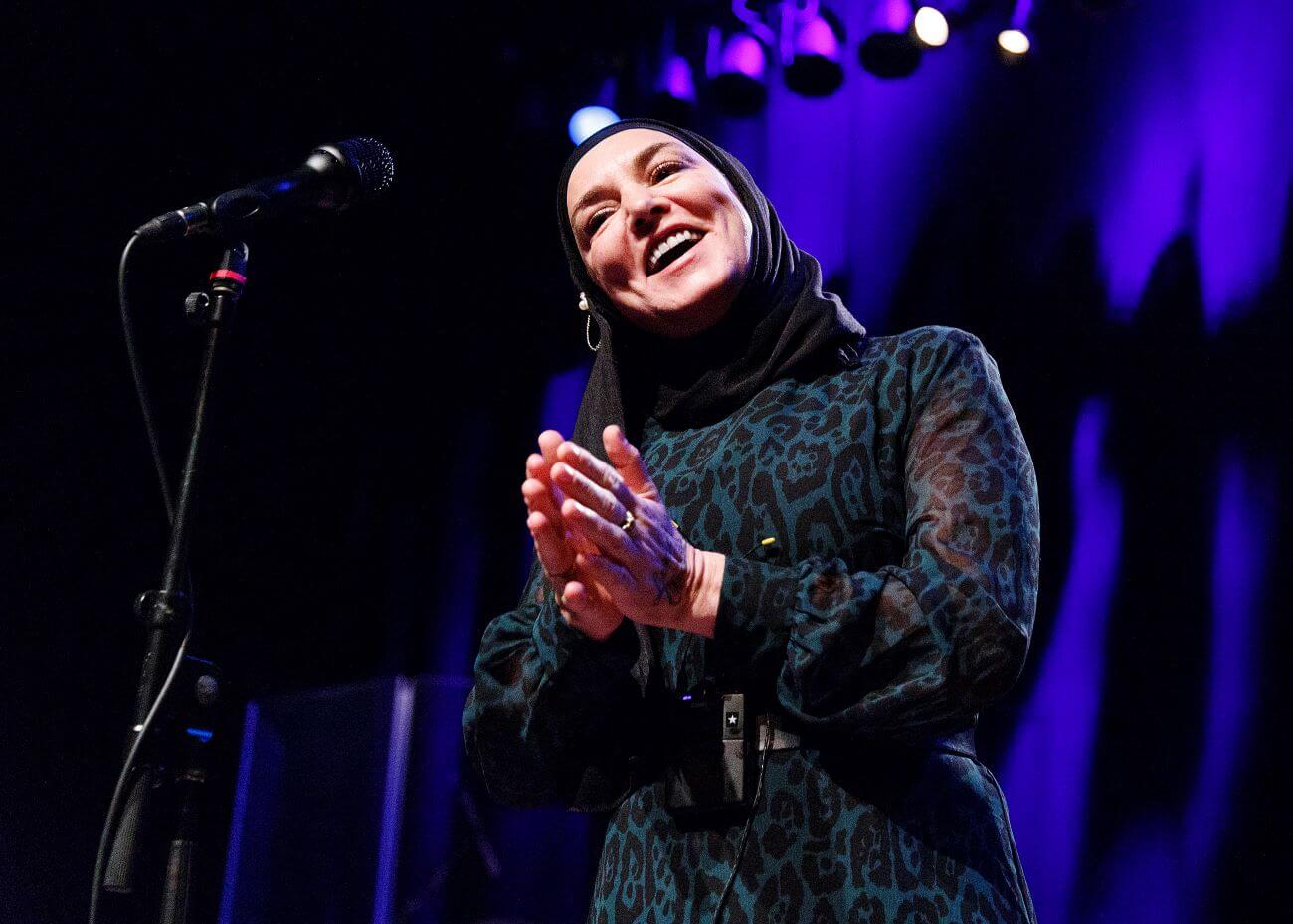 Sinead O'Connor wears a hijab and leopard print dress. She stands in front of a microphone and claps her hands together.