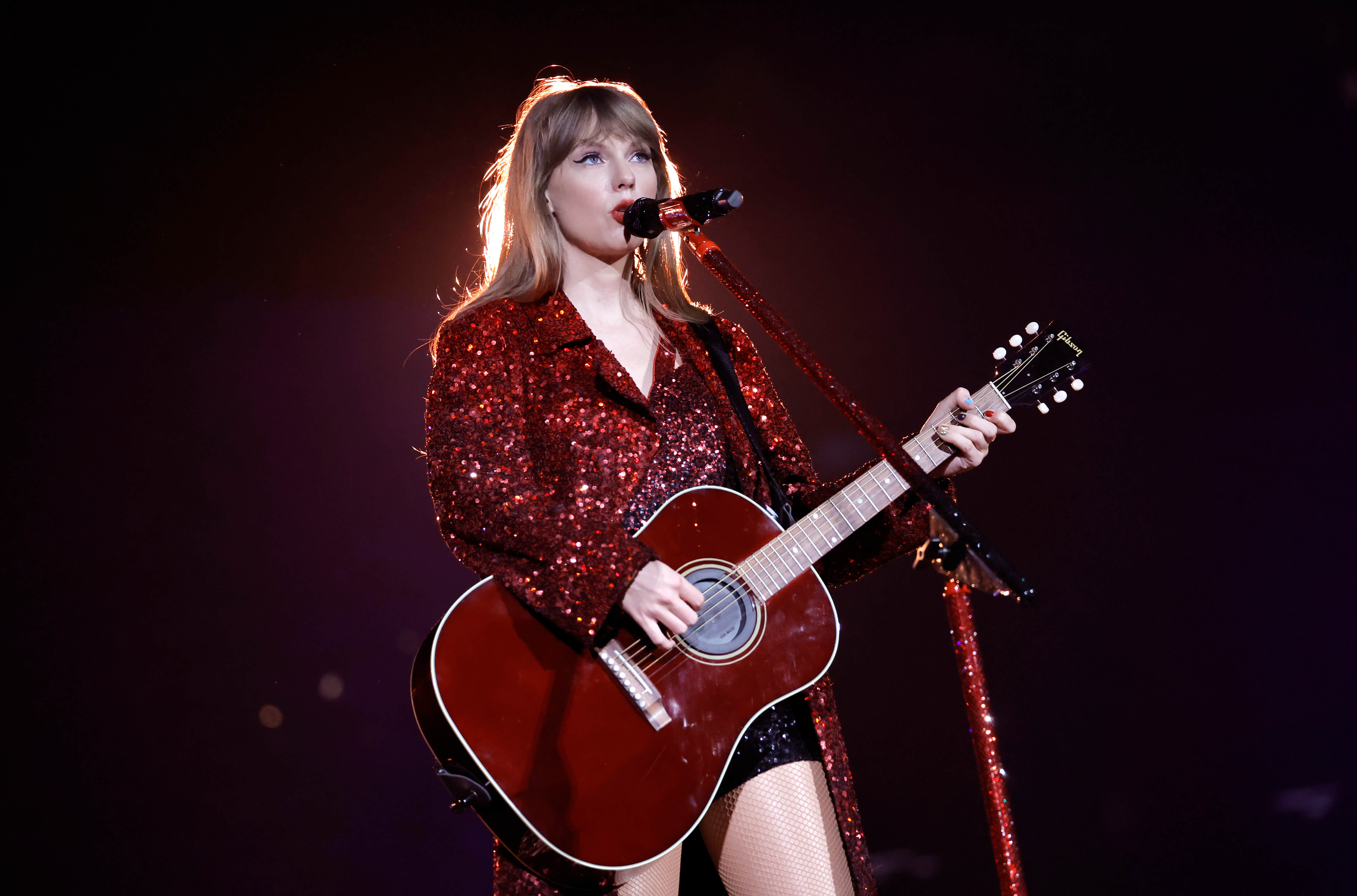 Taylor Swift playing guitar and singing into a microphone on stage.