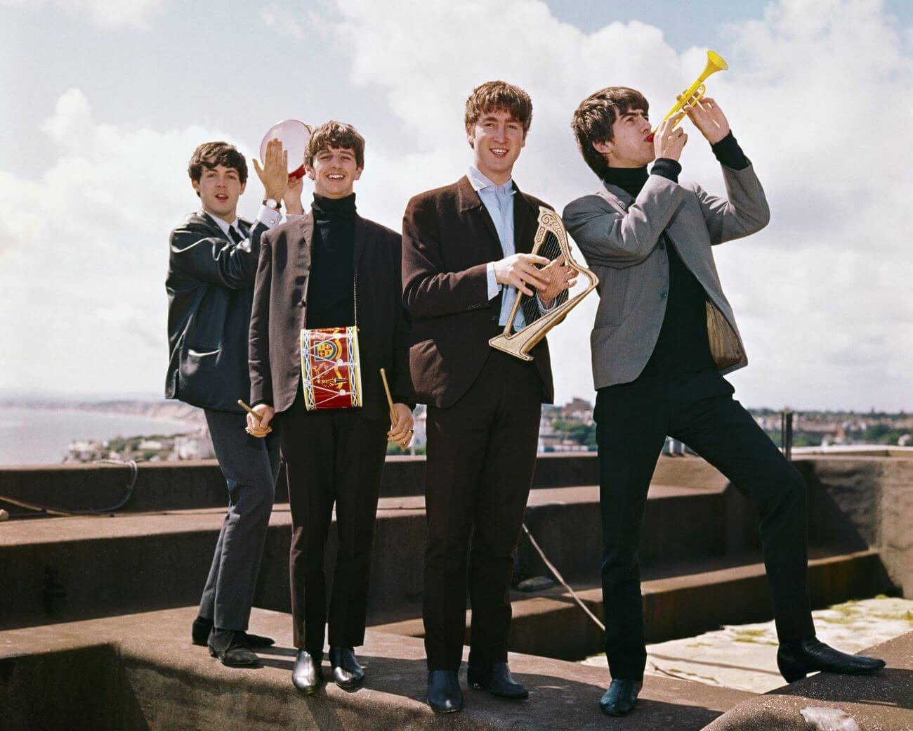The Beatles stand on a roof holding small instruments.